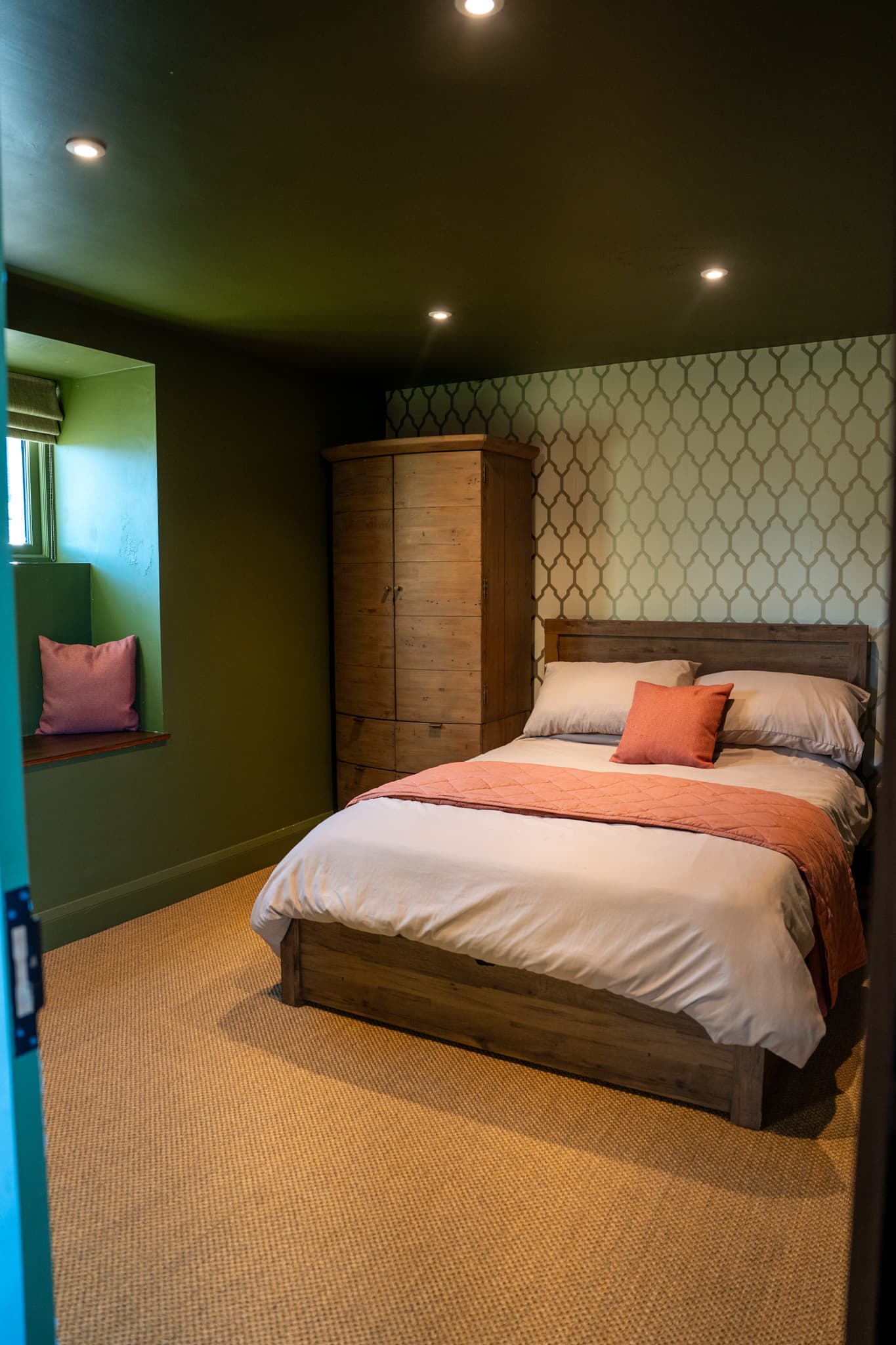 A ground-floor double bedroom with deep green walls and a patterned feature wall behind the bed. The wooden bed is made up in white linen with a coral throw and cushion, a tall pine wardrobe stands alongside, and a pink cushion sits in the deep window recess.