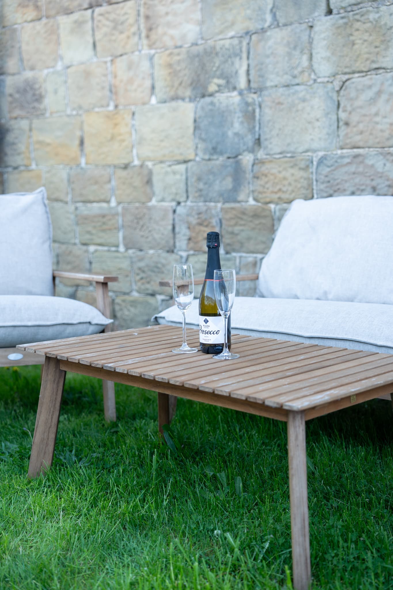 Teak garden furniture with thick cream cushions set on the grass against the barn’s honey-coloured stone wall, with a bottle of prosecco and two glasses on the slatted low table.