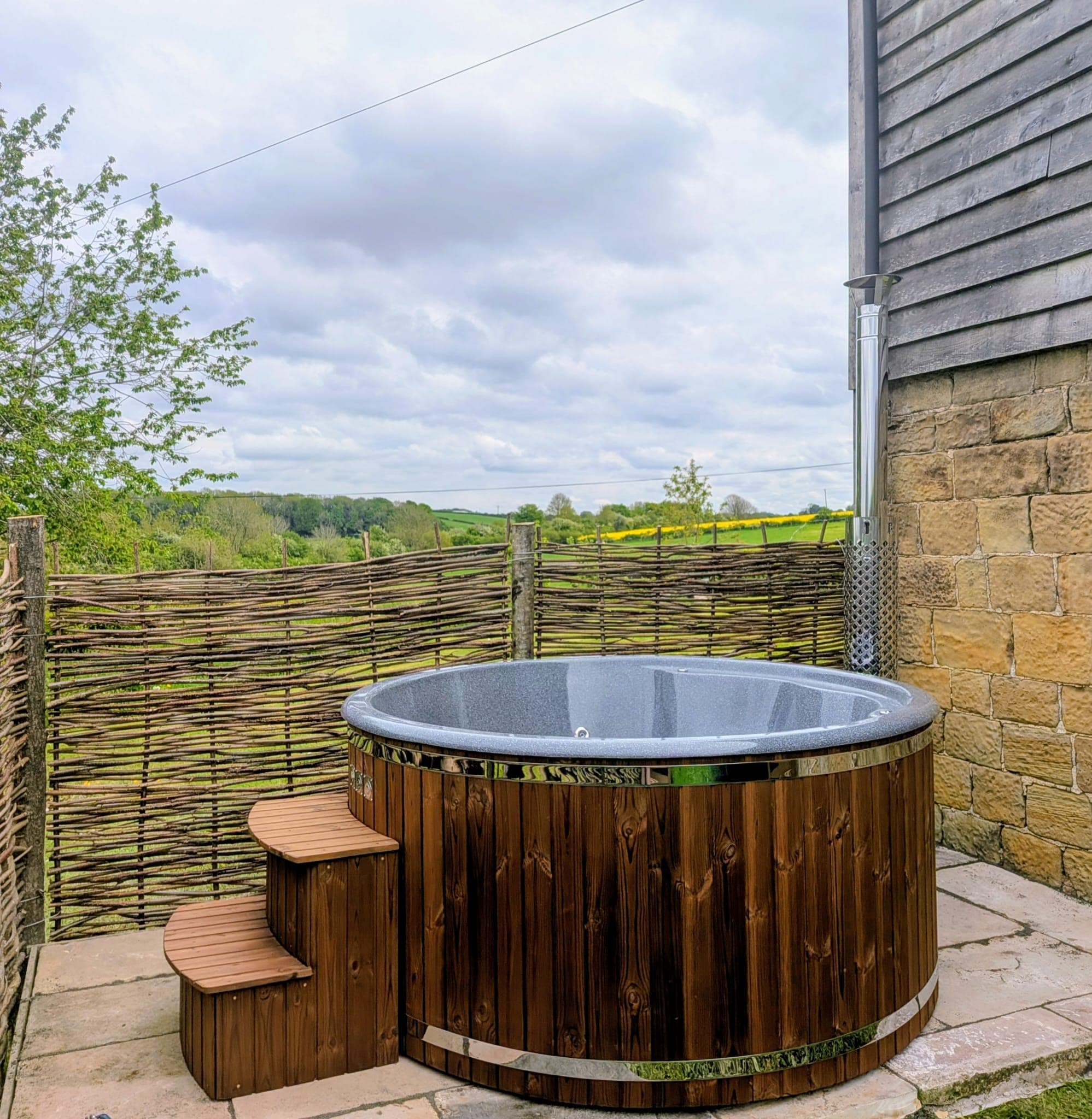 The wood-fired hot tub on the flagstone patio beside the barn, clad in dark timber with a stainless steel rim and wooden steps. A woven willow screen shelters it, and rolling countryside and a field of yellow rape stretch away behind.